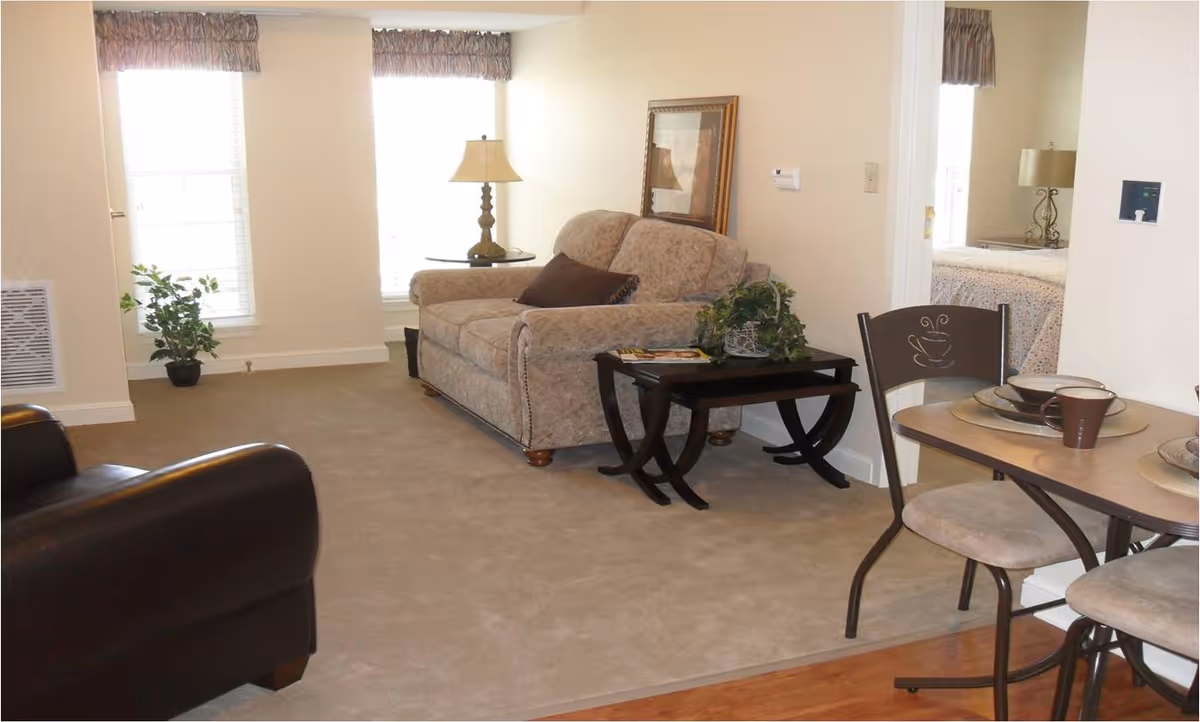 A cozy living area in an assisted living facility featuring a beige loveseat with a brown cushion, a dark wooden side table with a plant and magazines, a floor lamp, and two large windows with valances. Adjacent to the living area is a small dining table set with plates, bowls, and cups, and a glimpse of a bedroom with a bed and lamp is visible through an open doorway.