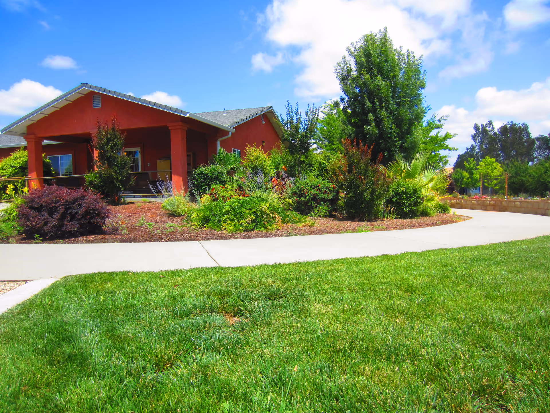 Single-story red building with a covered porch, landscaped shrubs, a curved walkway and green lawn under a blue sky.