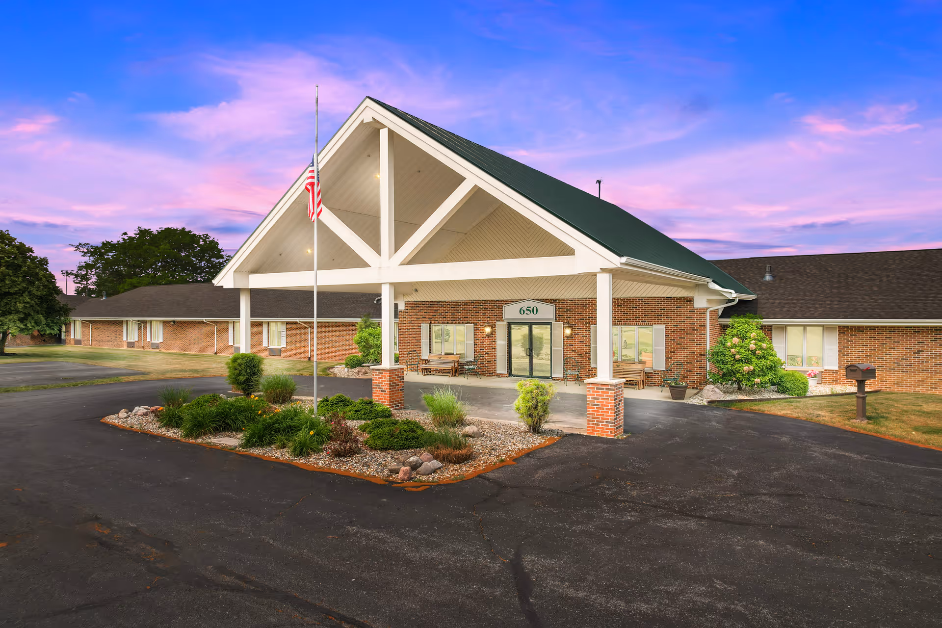 Exterior view of Vancrest of Payne senior living facility at sunset, showing a brick building with a green roof, a covered entrance with white pillars, an American flag on a flagpole, landscaped greenery, and a paved driveway.