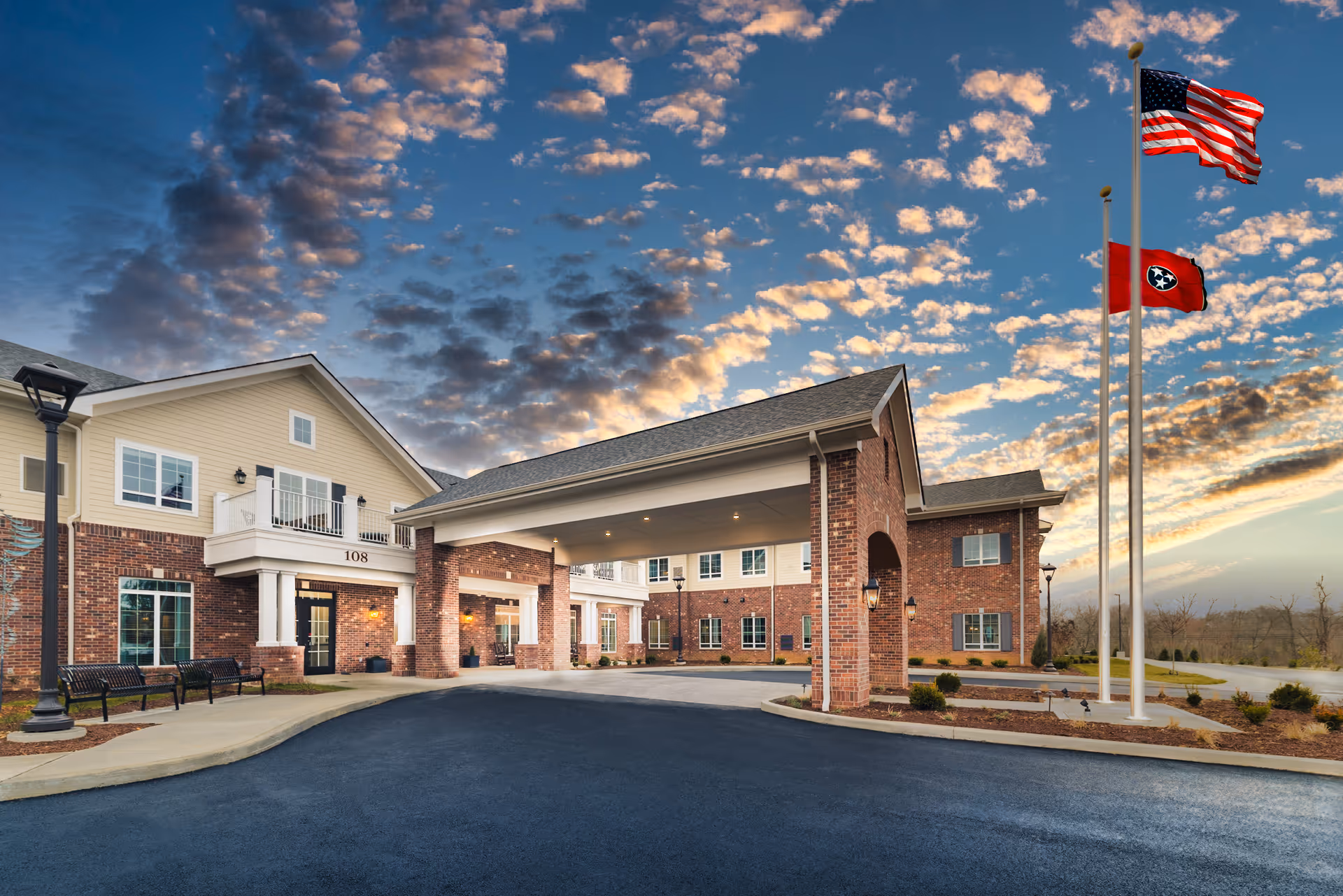 Brick senior living facility with porte-cochère entrance, flags, and curved driveway at dusk.