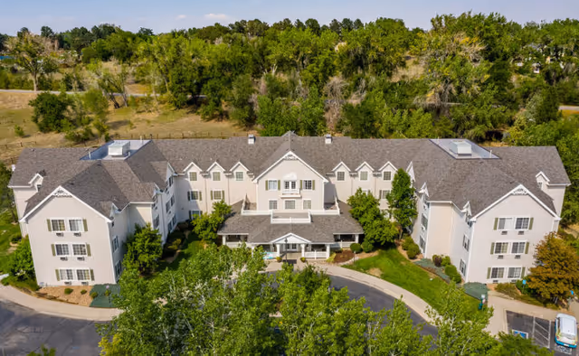 Aerial view of a large, three-story senior living facility building surrounded by trees and greenery. The building has a gray roof and light-colored exterior walls with multiple windows. There is a circular driveway and landscaped areas with bushes and trees in front of the entrance.
