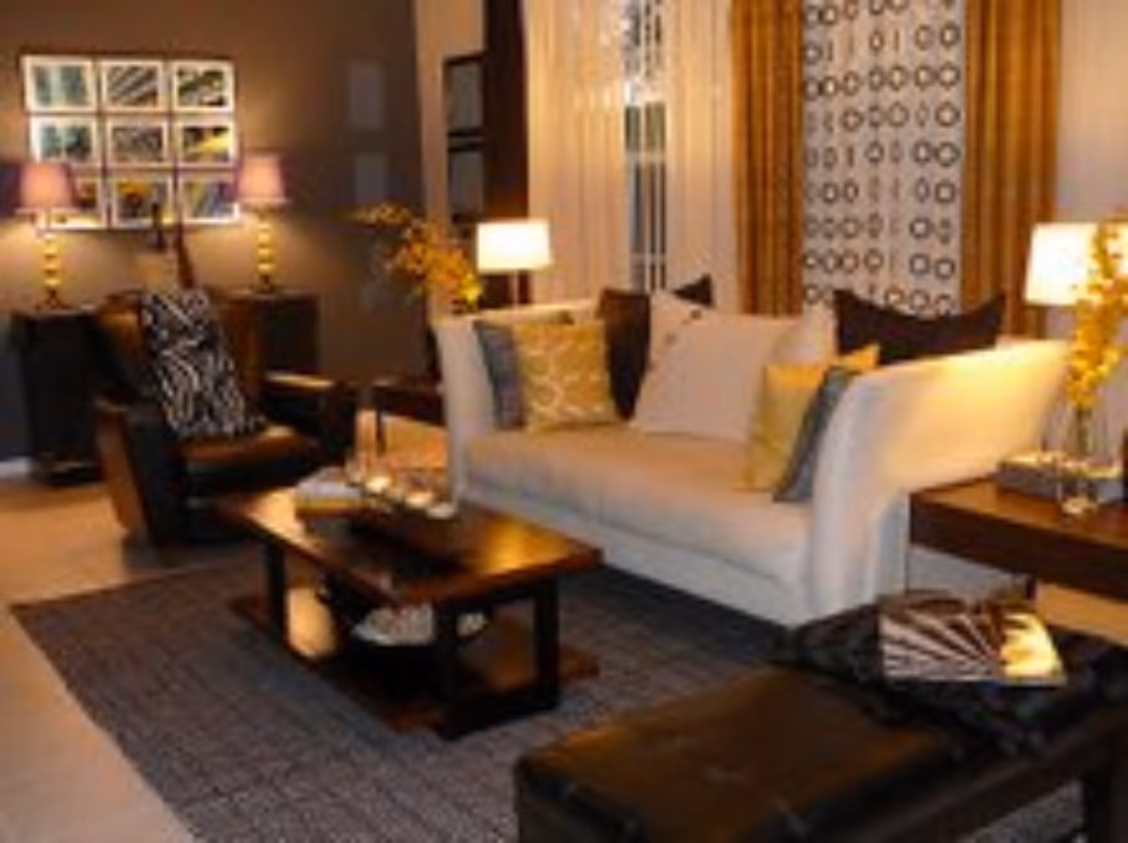 A cozy living room with a white sofa adorned with multiple cushions, a dark wooden coffee table with decorative items, a black armchair with a patterned cushion, and a matching ottoman. The room is warmly lit by table lamps on side tables, with framed pictures on the wall and patterned curtains on the window.