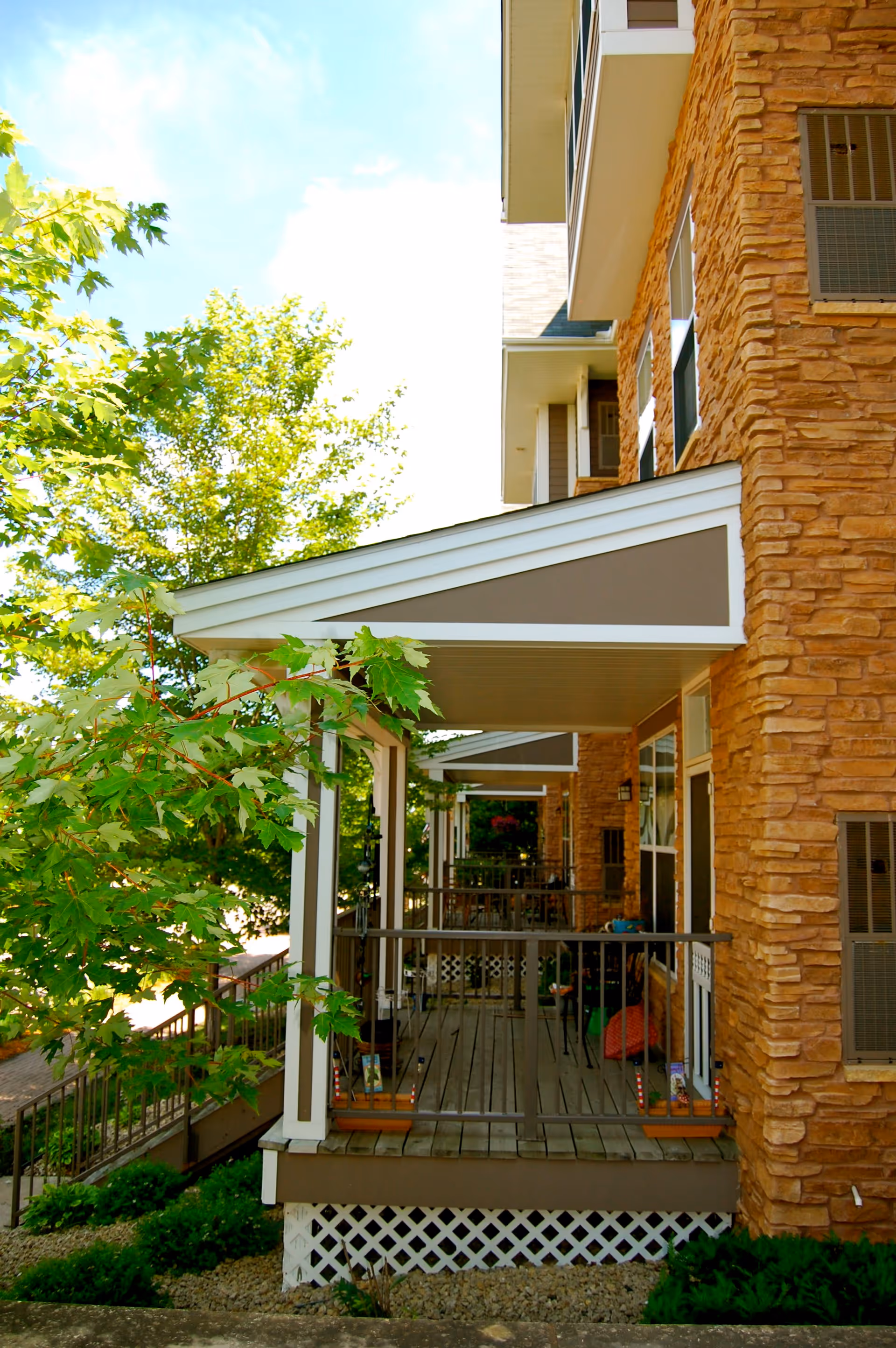 View of a covered porch area attached to a building with stone exterior walls, surrounded by green trees and shrubs under a bright sky.
