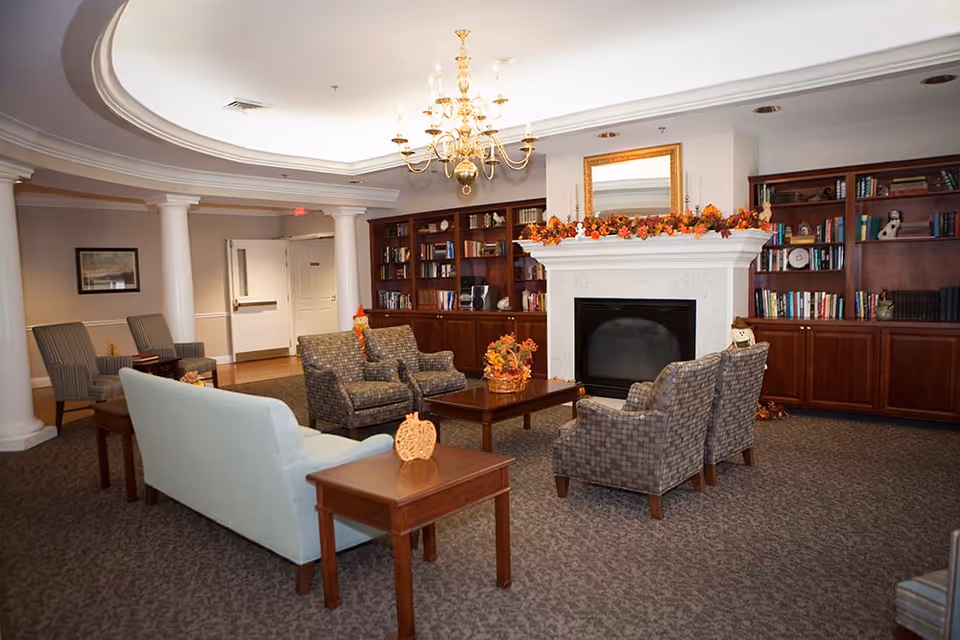 A cozy living room area with a white fireplace decorated with autumn-themed garlands and a mirror above it. Surrounding the fireplace are wooden bookshelves filled with books and decorative items. The seating includes a light blue sofa, four patterned armchairs, and two striped chairs, arranged around a wooden coffee table with a fall-themed centerpiece. The room features a chandelier, carpeted floor, and white columns near the entrance.