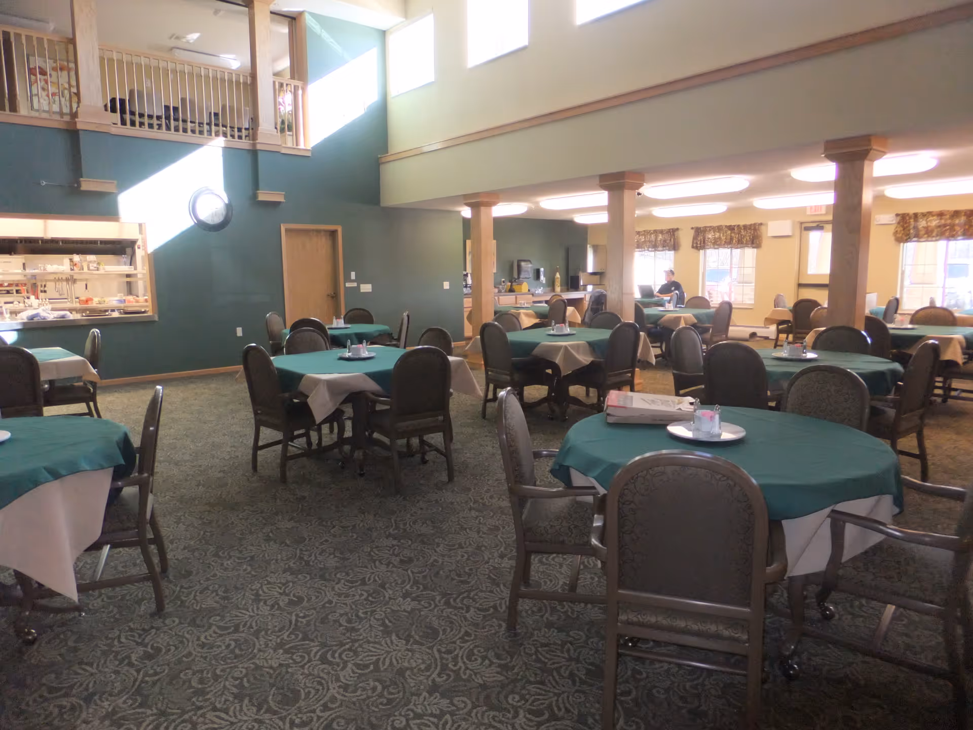 Spacious dining room with round tables covered in green tablecloths and chairs in a senior living facility.