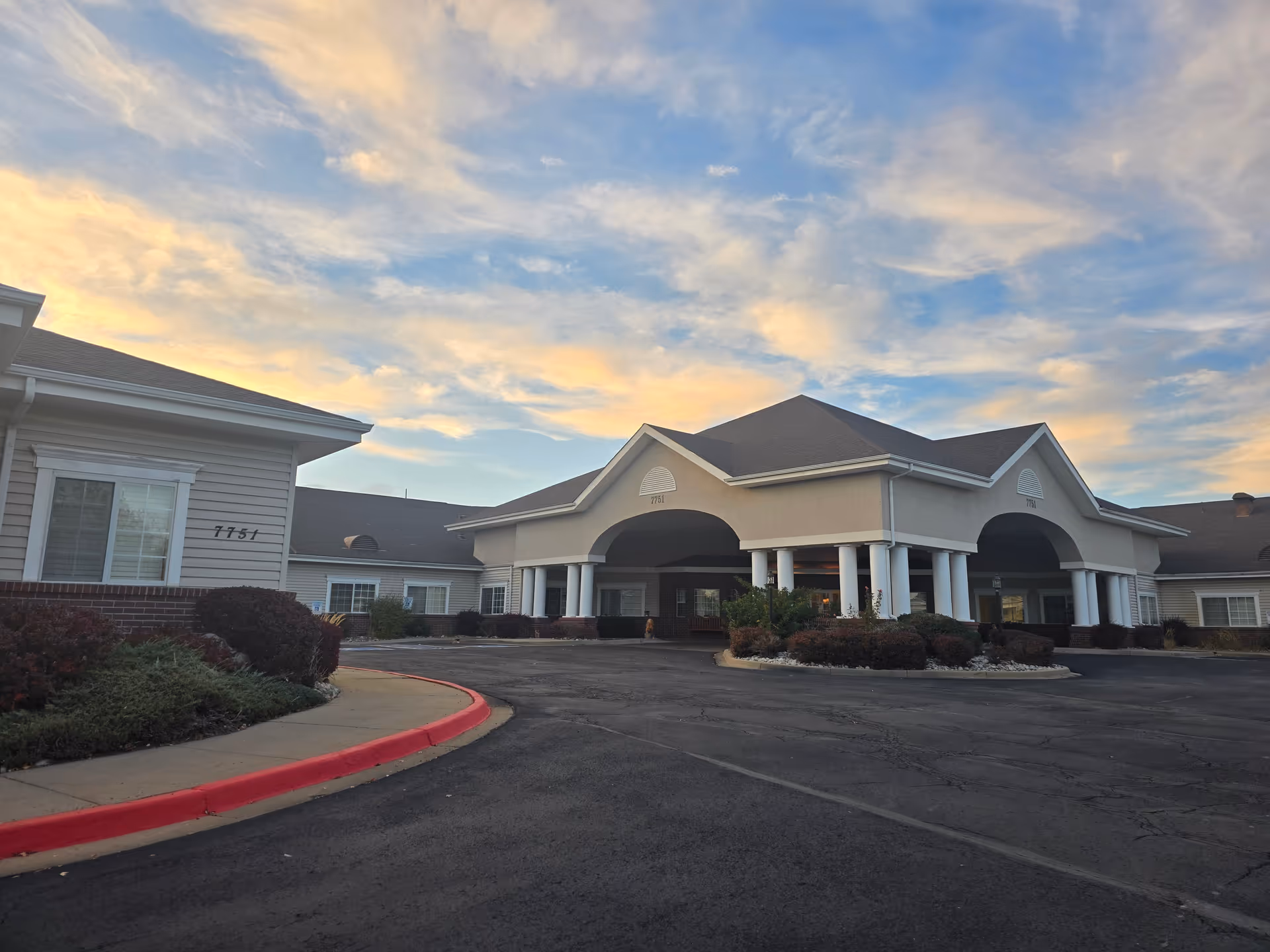 Exterior view of the Life Care Center of Westminster building at sunset, showing the entrance with a covered driveway, columns, and surrounding landscaping with bushes and a curved sidewalk.