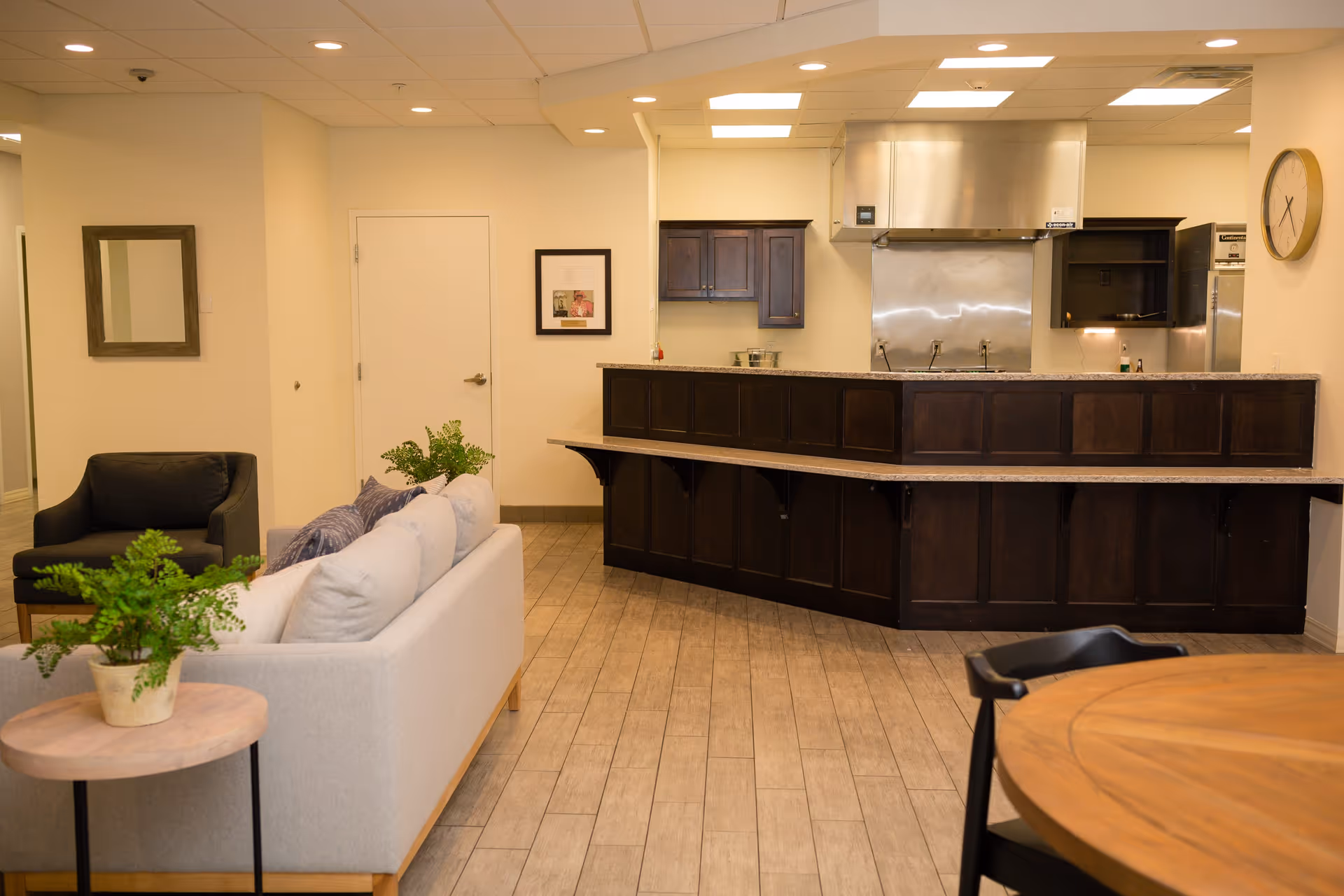Interior view of a memory care facility showing a cozy seating area with a light gray sofa, a black armchair, and a small round wooden table with a potted plant. In the background, there is a kitchen area with dark wood cabinetry, a stainless steel range hood, and a countertop bar. The room has light-colored walls, wood-look tile flooring, a wall clock, and framed artwork.