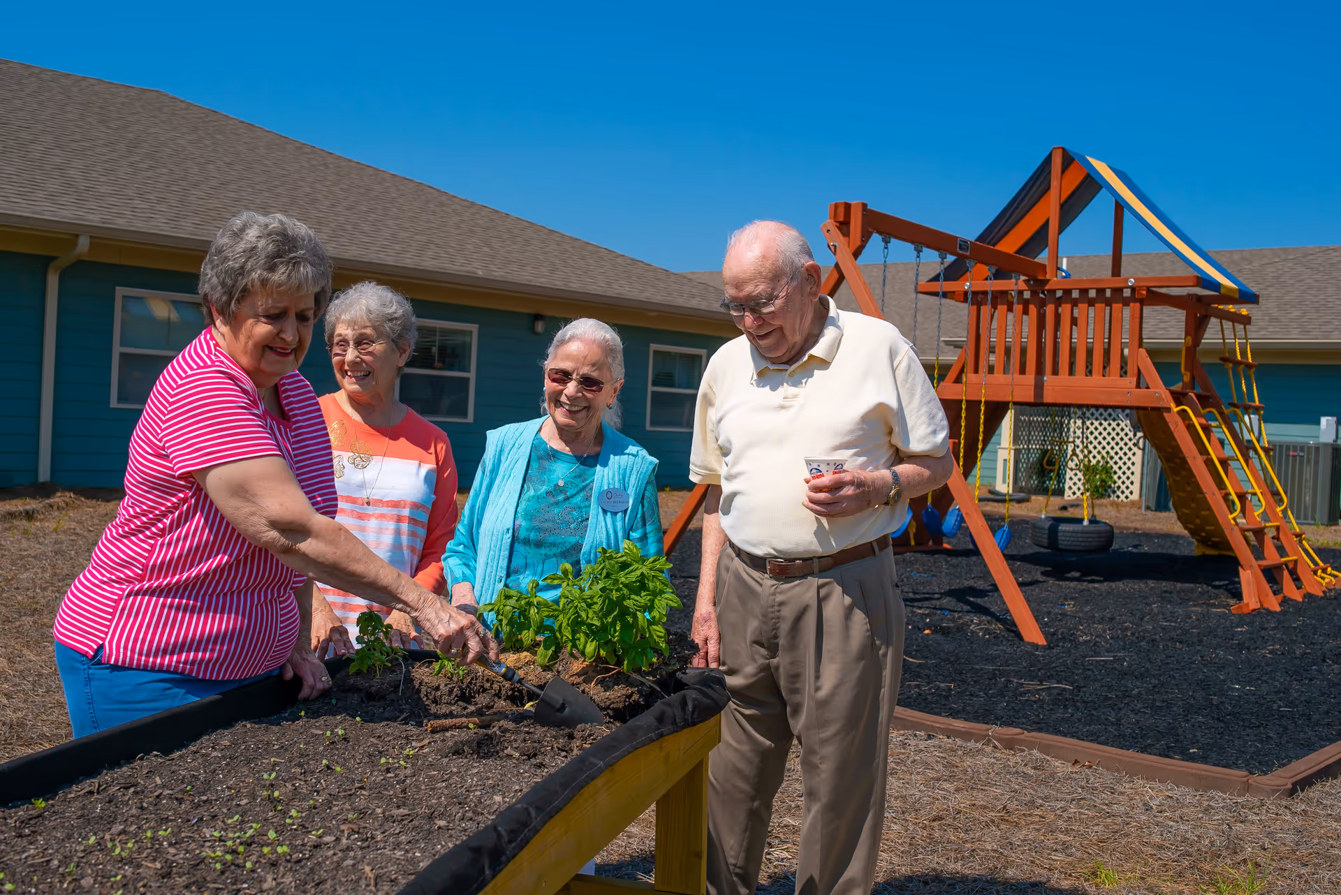 Four elderly people, three women and one man, are gathered around a raised garden bed outdoors. One woman is tending to the plants with a small gardening tool while the others watch and smile. In the background, there is a playground structure with swings and a slide, and a building with blue siding and windows under a clear blue sky.