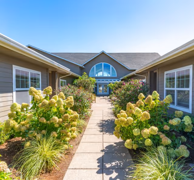 Wide walkway flanked by flowering shrubs leads to the front entrance of a senior living center.