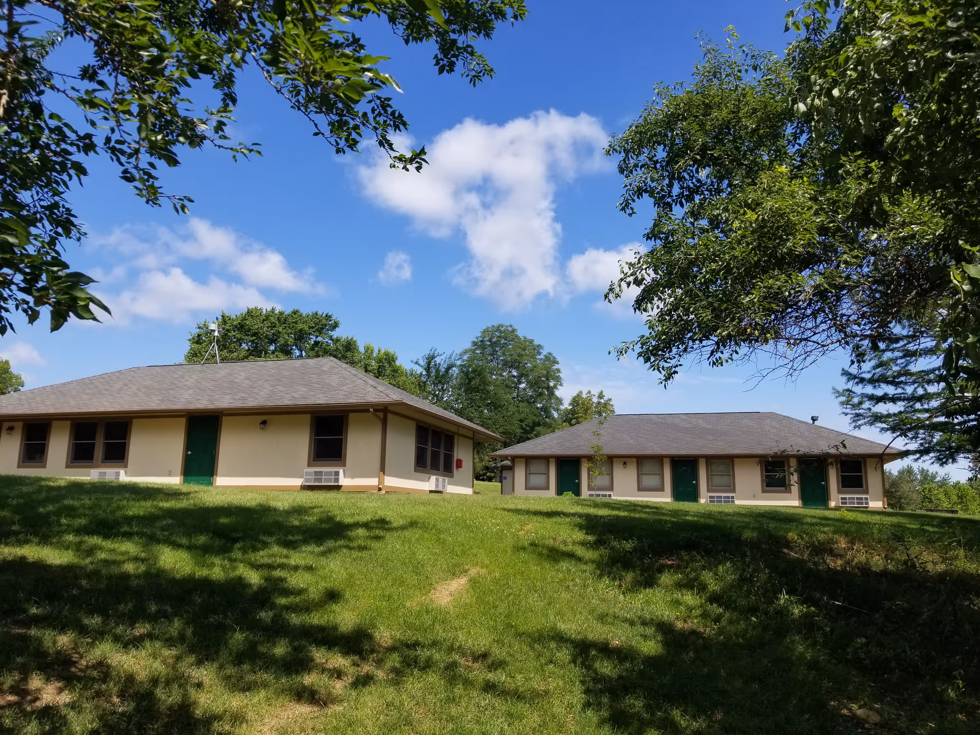 Two beige, single-story buildings with green doors sit on a grassy hill under a blue sky.