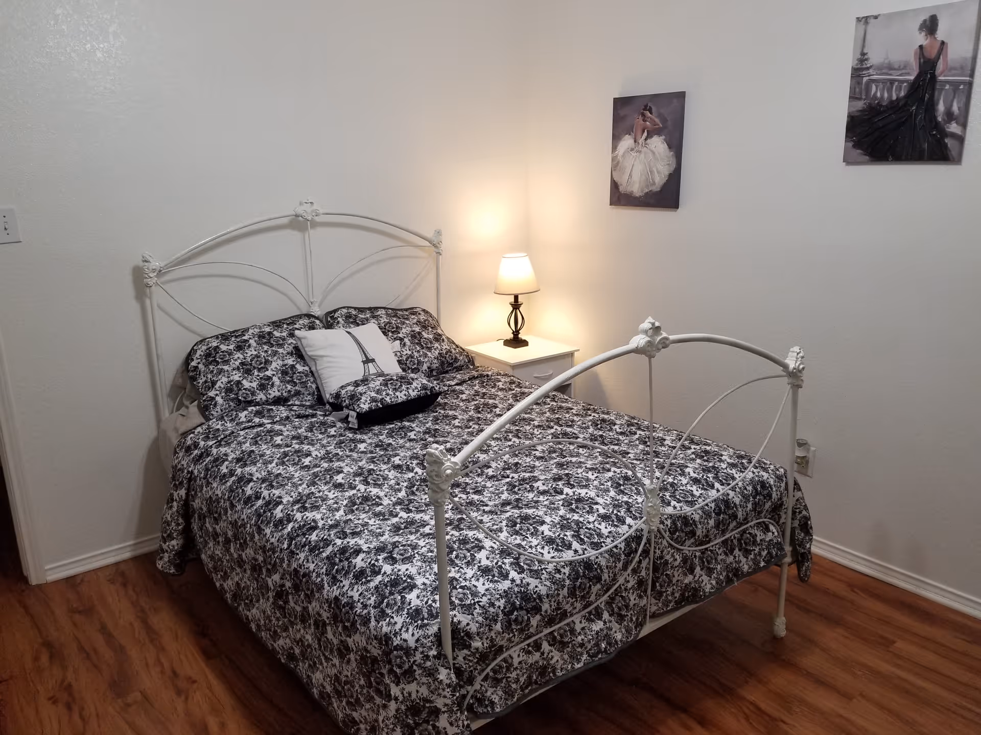 White metal bed with floral bedding, nightstand, lamp, and wall art in a bedroom.