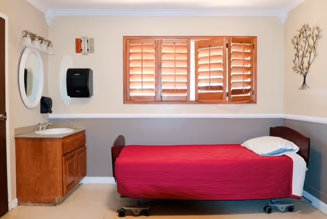 A small senior living bedroom with a red bed, window blinds, and a sink cabinet.