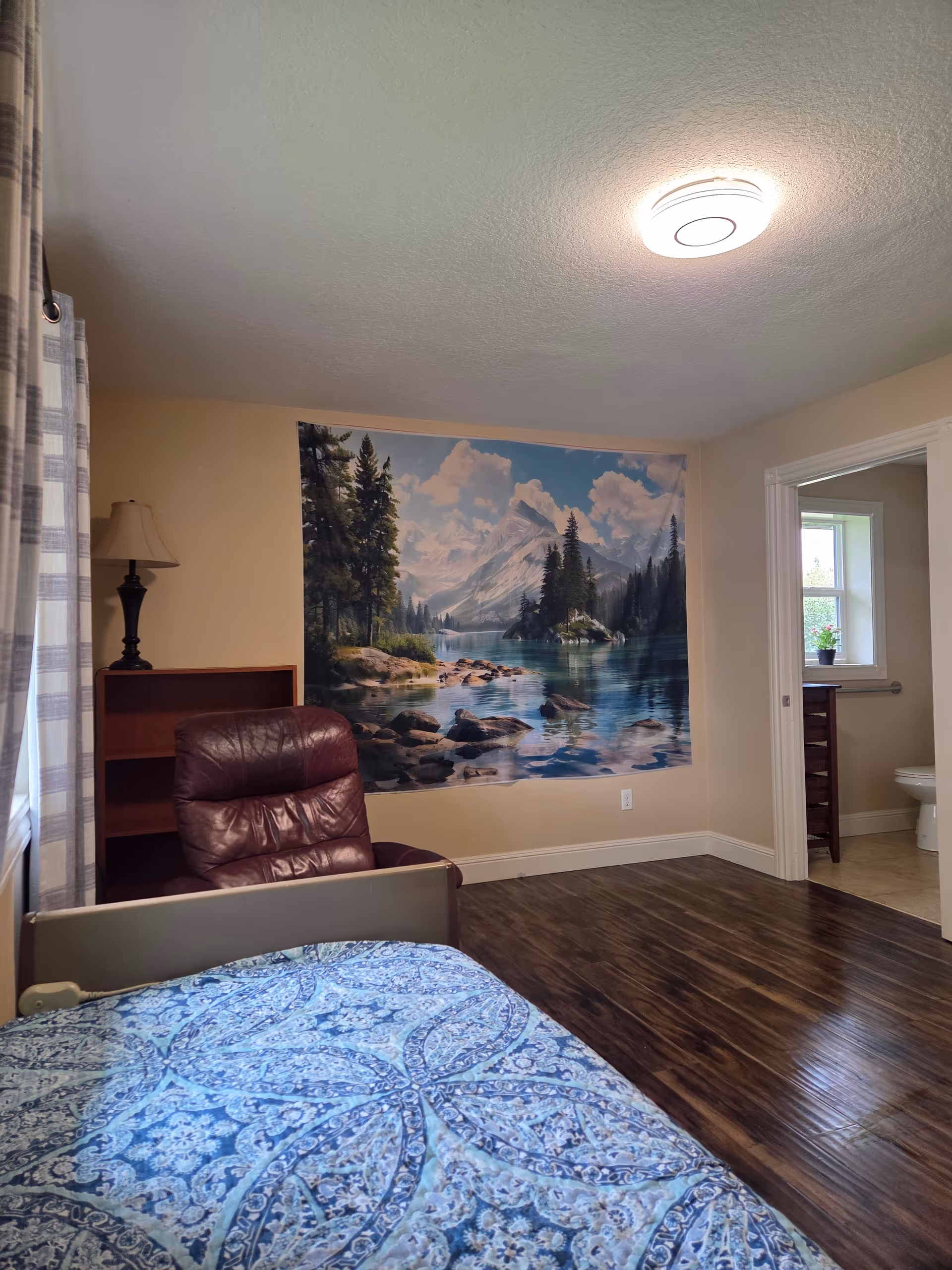 Bedroom in a senior home featuring a bed, burgundy chair, and mountain mural.