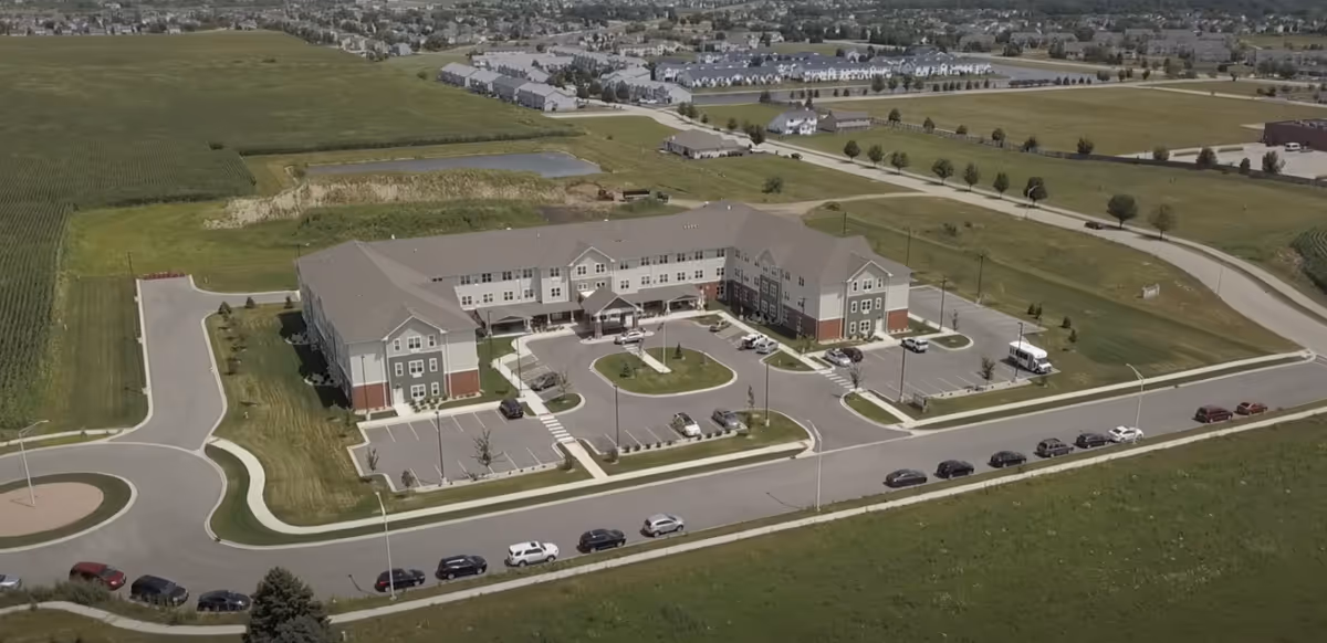 Aerial view of a large three-story senior living facility with parking lots, driveways, and surrounding suburban landscape.