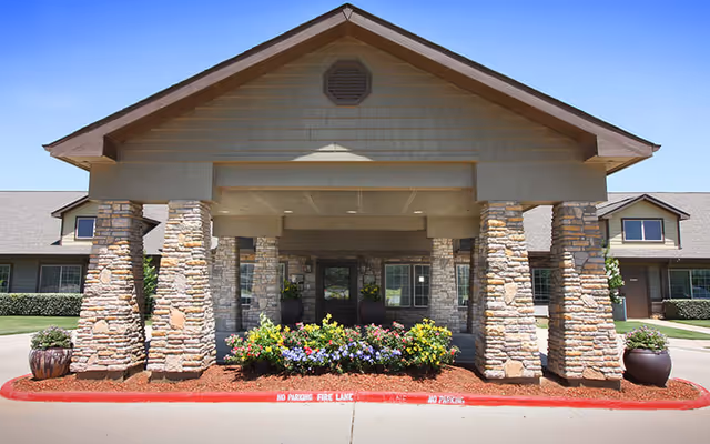 Covered main entrance with a gabled portico supported by stone columns, potted plants, and flower beds in front of a single-story building.