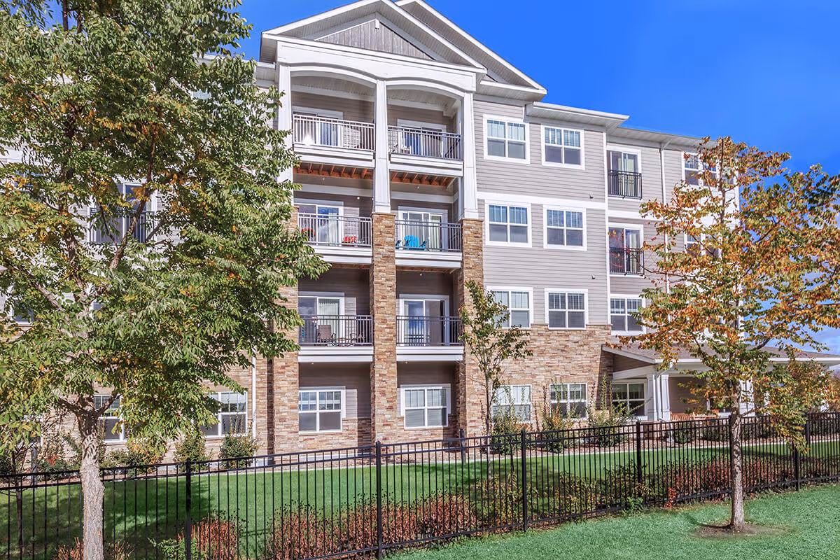 Front exterior of a multi-story assisted living building with balconies, landscaping, and a fenced lawn.