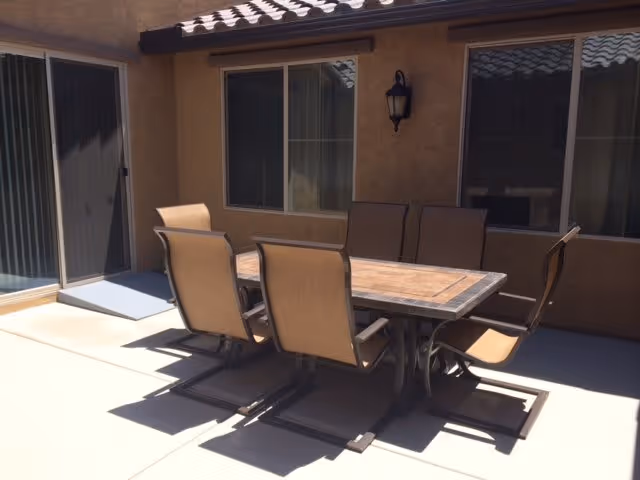 Outdoor patio area with a rectangular table and six cushioned chairs arranged around it. The patio is adjacent to a building with beige walls, two windows, and a sliding glass door. The area is well-lit by sunlight.