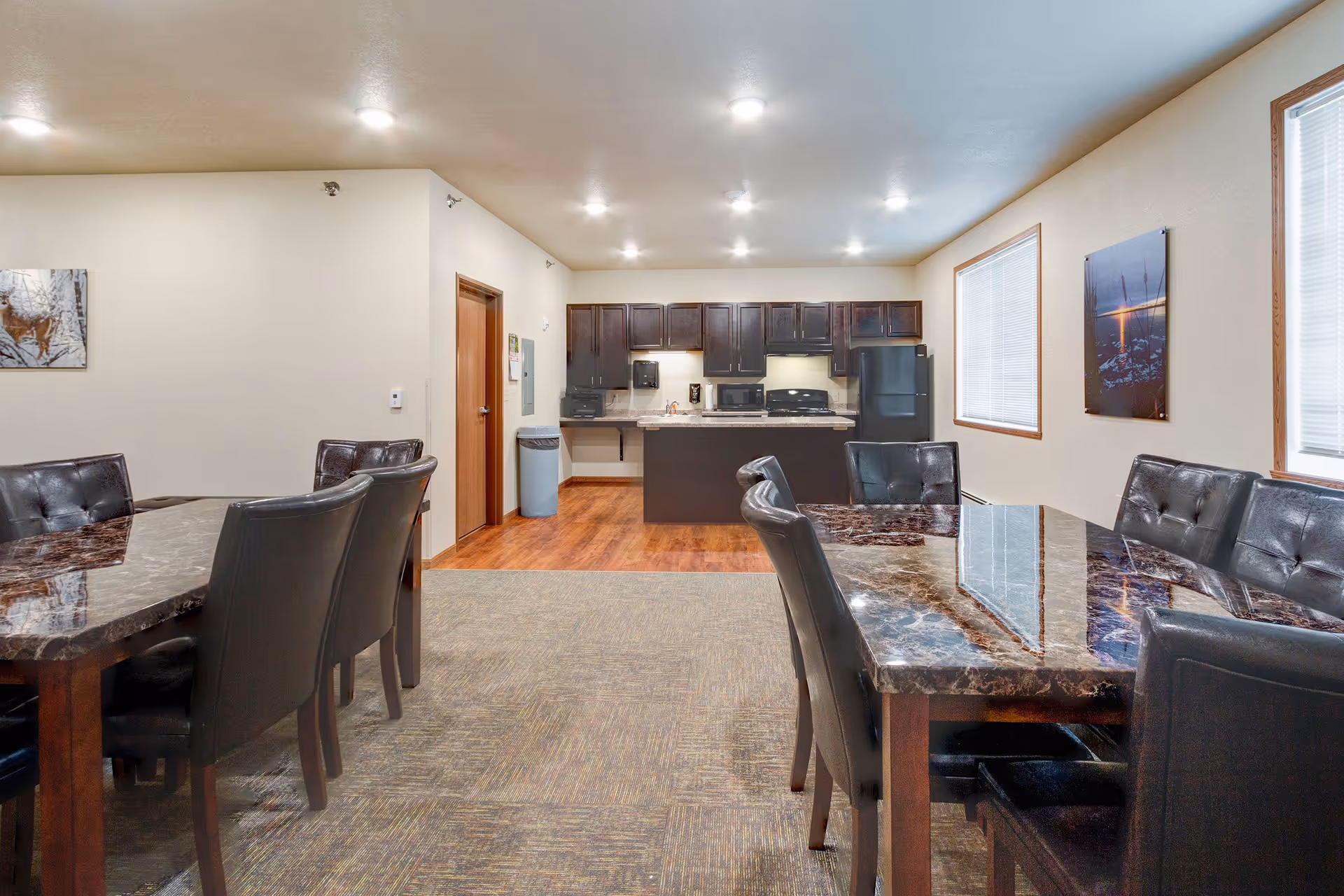 Interior view of a communal dining and kitchen area with two marble-top dining tables surrounded by black cushioned chairs. The kitchen area features dark wood cabinets, a black refrigerator, microwave, stove, and a countertop. The room has beige walls, carpet and wood flooring, and two windows with blinds. There are framed pictures on the walls.