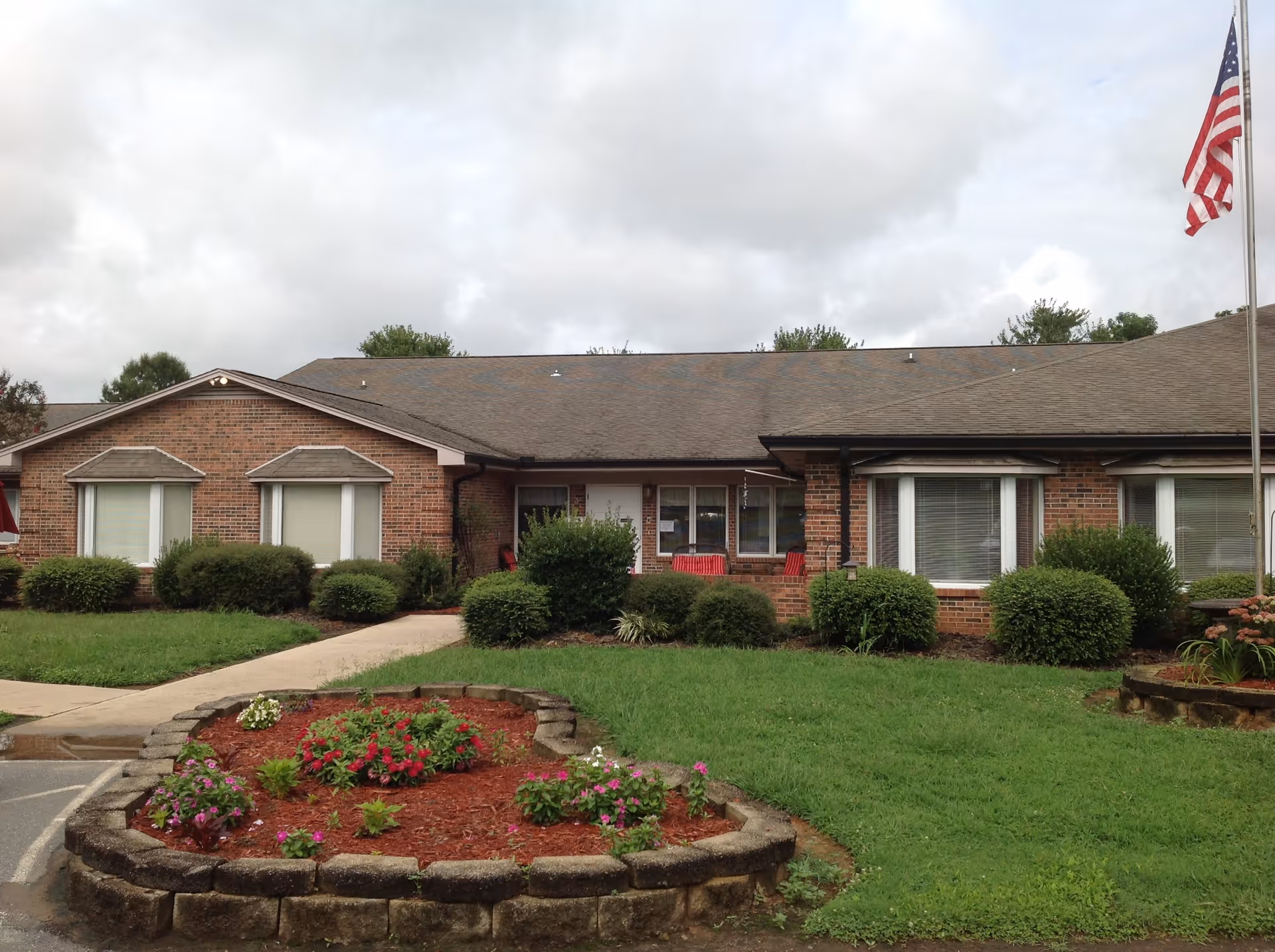 Front exterior view of a single-story brick building with multiple windows, a small porch with red chairs, well-maintained bushes, a flower bed with colorful flowers, green lawn, and an American flag on a flagpole to the right.
