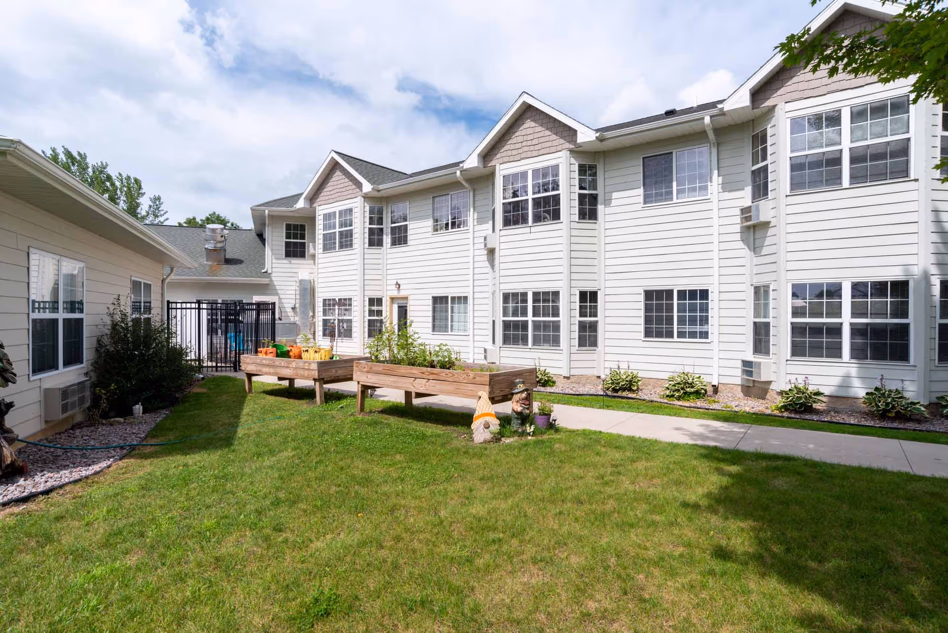 Two-story white senior living building with a grassy courtyard, raised garden beds and a concrete walkway.
