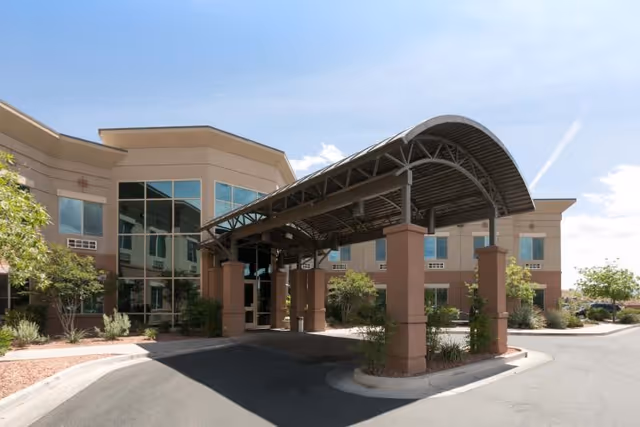 Exterior view of Coral Desert Rehabilitation And Care facility showing a covered entrance with a curved metal roof supported by columns, surrounded by a paved driveway and landscaping with trees and shrubs under a clear blue sky.