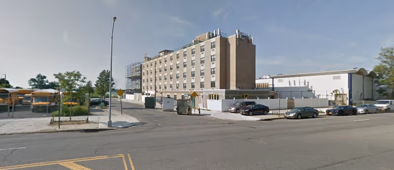 A multi-story brick institutional building with parked cars and school buses along the street in front.
