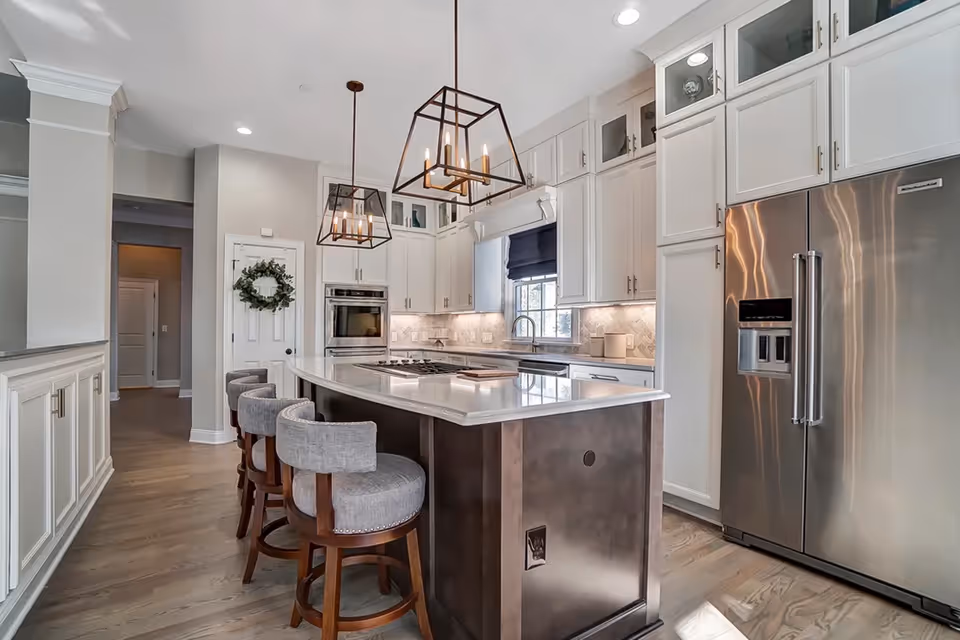 A modern white kitchen with an island, pendant lights, and stainless steel appliances.