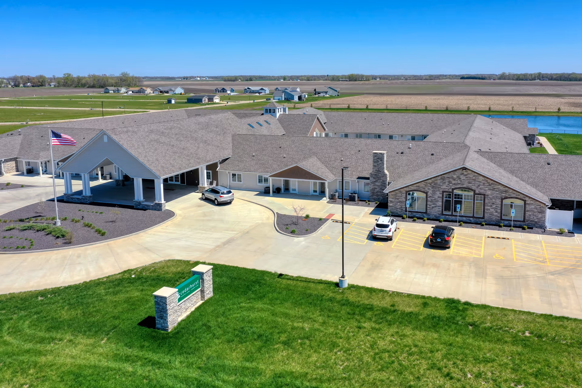 Aerial view of Cedarhurst Senior Living of Bethalto showing a large single-story building with a covered entrance, a parking lot with a few cars, an American flag on a flagpole, and surrounding green grass and farmland under a clear blue sky.