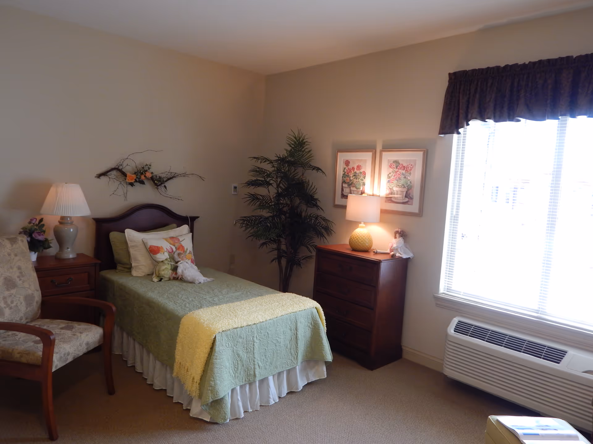 A neatly furnished bedroom with a single bed dressed in green bedding, a wooden dresser and lamp, an armchair, potted plant, and a large window with blinds.