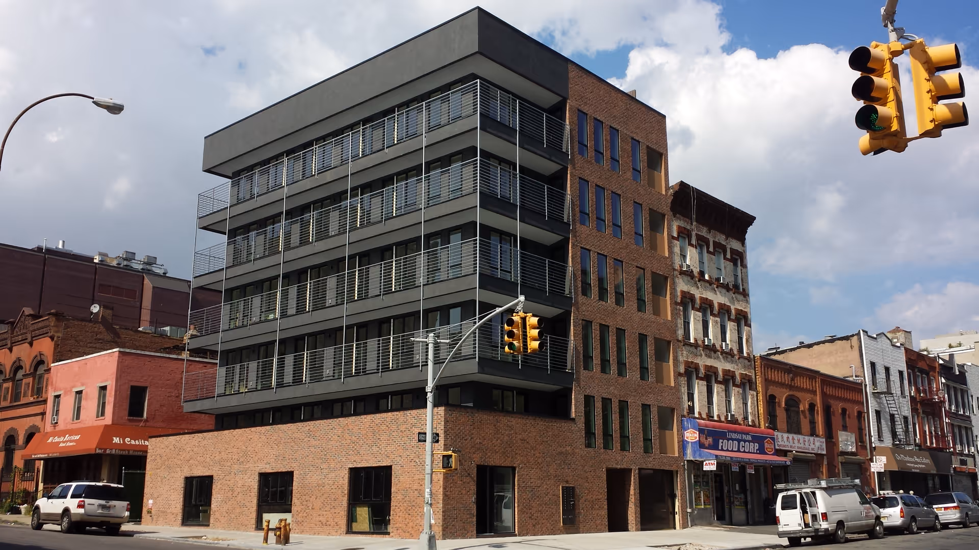 Corner street view of a modern brick-and-concrete building with balconies and adjacent shops.