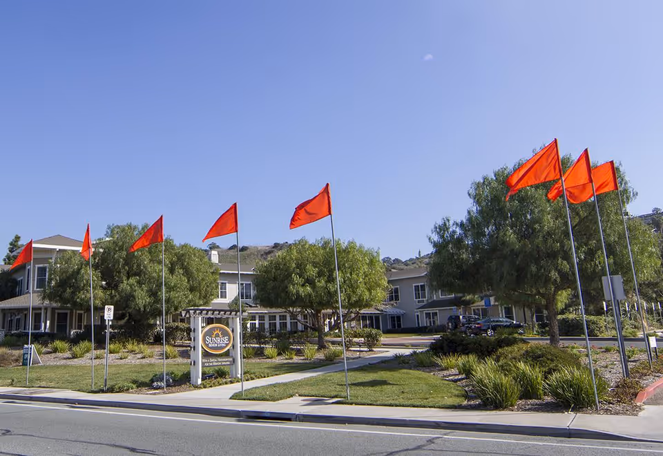 Exterior view of a senior living facility with a two-story building surrounded by trees and landscaping. Several red flags on poles line the sidewalk in front of the building. A sign near the entrance reads 'Sunrise Senior Living'. The sky is clear and blue.
