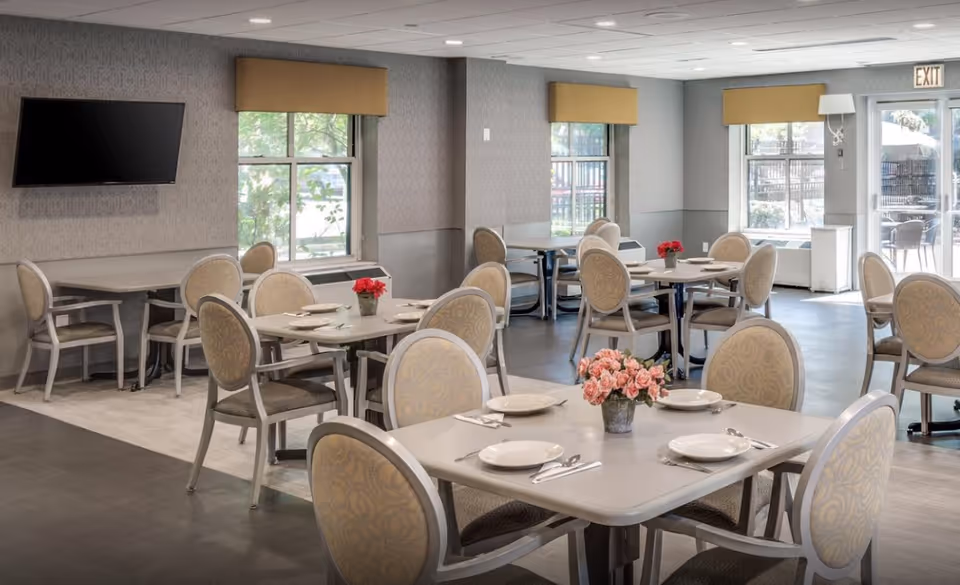 A bright dining room with round tables, floral centerpieces, and cushioned chairs.