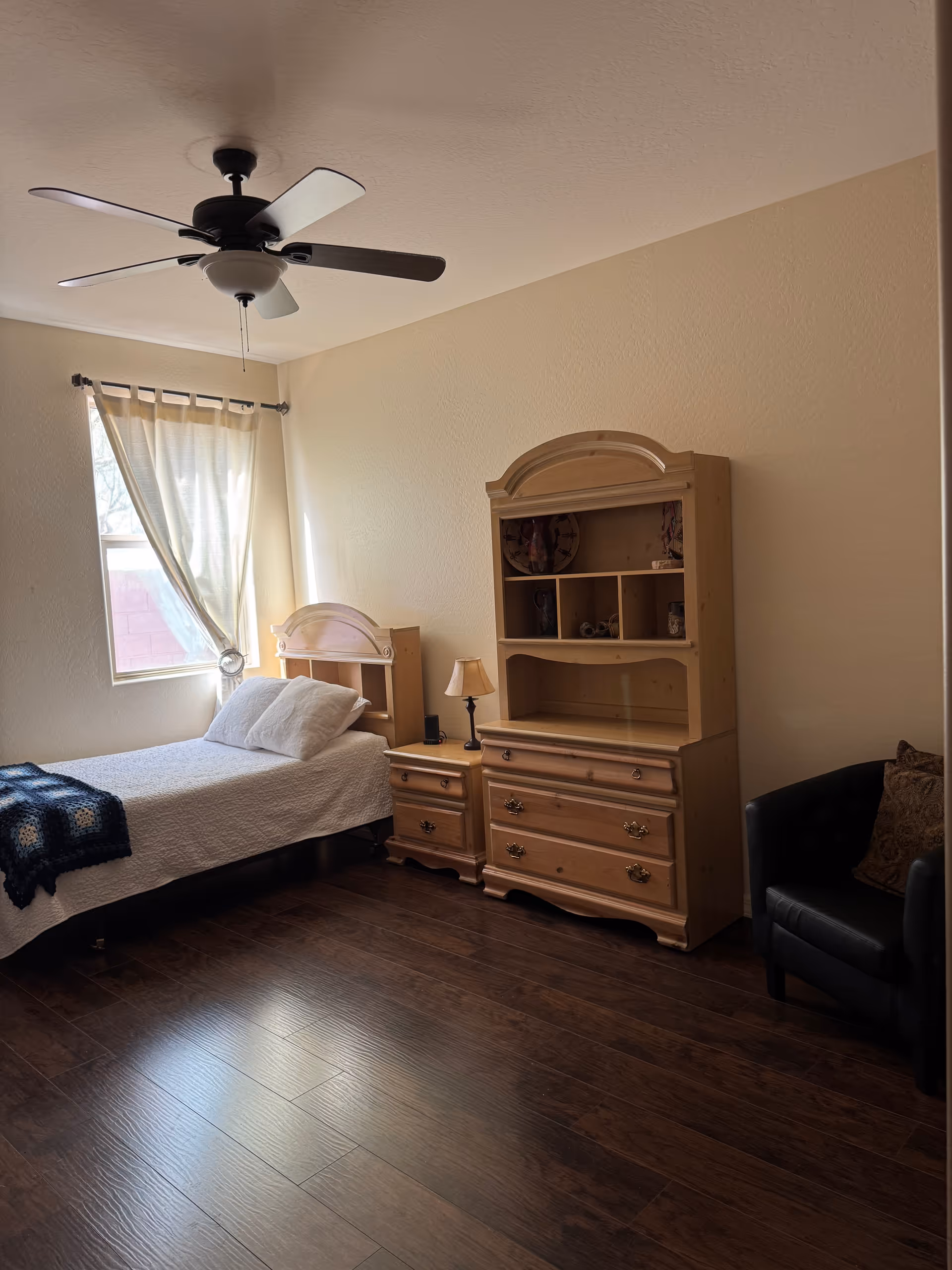 A cozy bedroom with a single bed, dresser with hutch, nightstand, ceiling fan, and window.