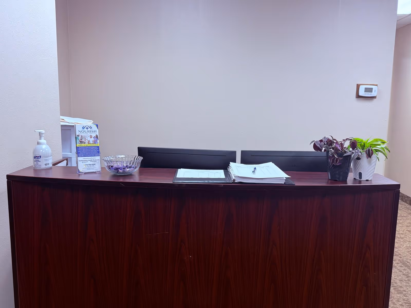 A dark wood reception desk with sanitizer, brochures, a binder, and potted plants in a lobby.