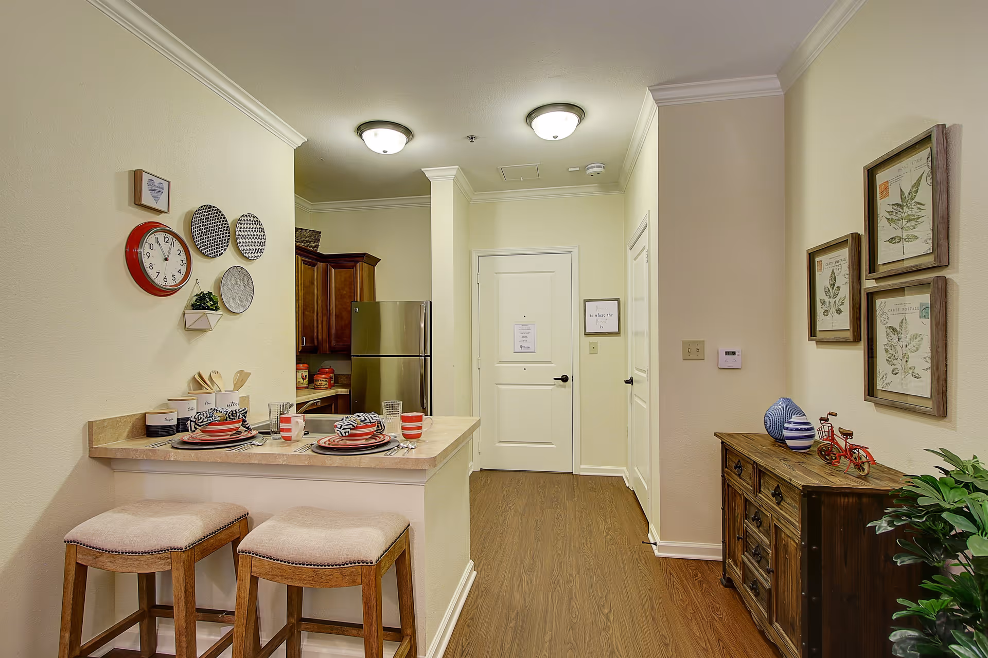 Interior view of a small kitchen and dining area in a senior living facility. The kitchen features wooden cabinets, a stainless steel refrigerator, and a countertop with two cushioned stools. The countertop is set with plates, bowls, and cups. The walls are decorated with a clock, decorative plates, and framed botanical prints. There is a wooden sideboard with decorative items and a plant on the right side. The floor is wooden, and the walls are painted light beige.