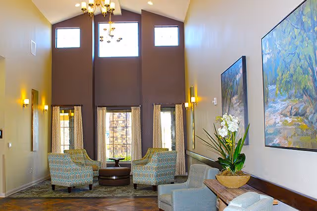 Vaulted communal lounge with armchairs, a chandelier, tall windows, artwork and a potted plant.