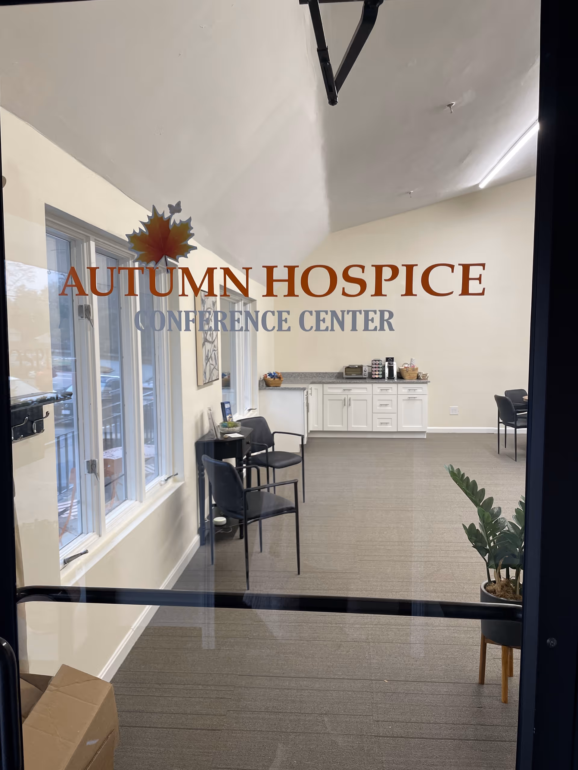 Glass door shows Autumn Hospice Conference Center with kitchenette, chairs, and large windows inside.