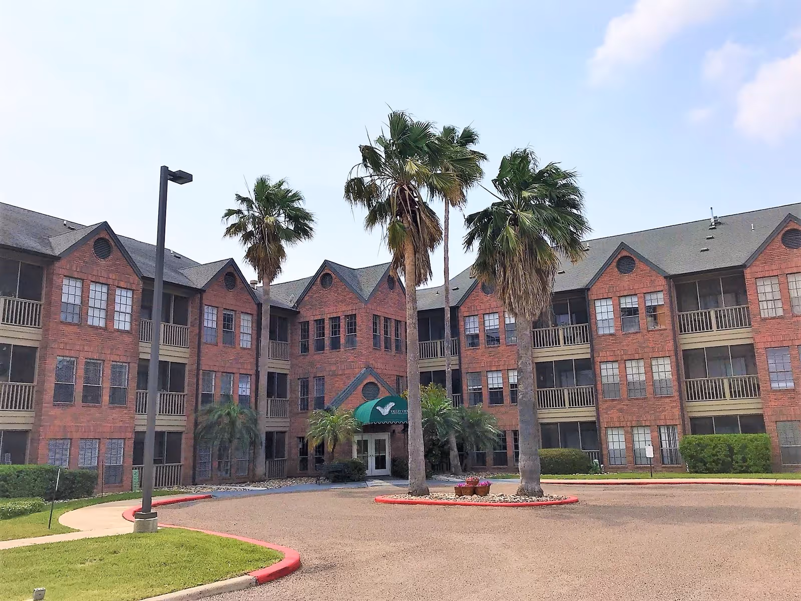 Front exterior view of a three-story brick building with multiple windows and balconies, surrounded by palm trees and a circular driveway with a green awning entrance.