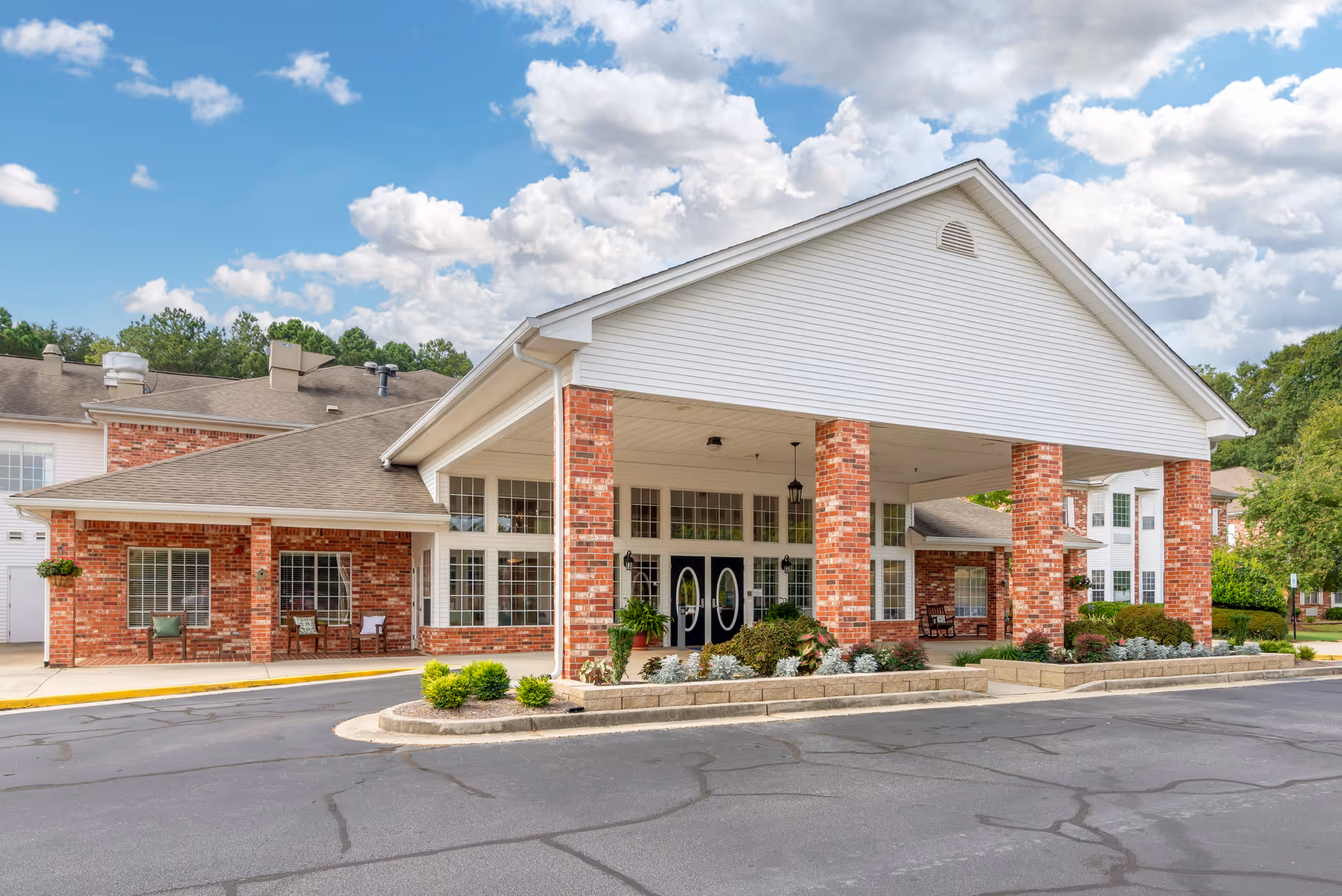 Exterior view of a senior living facility with a covered entrance supported by brick columns, large windows, and a landscaped area with shrubs and plants under a partly cloudy sky.