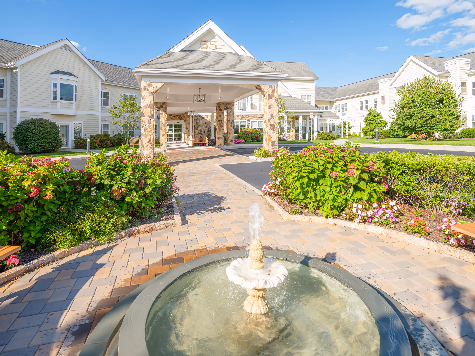 Front entrance of a light-colored senior living facility with a fountain in the foreground, landscaped flowerbeds, and a covered porte-cochere.