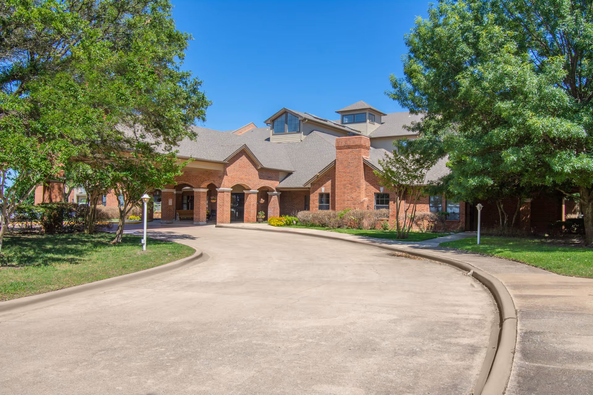 Front entrance of a brick senior living facility with a covered portico, curved driveway, and trees.