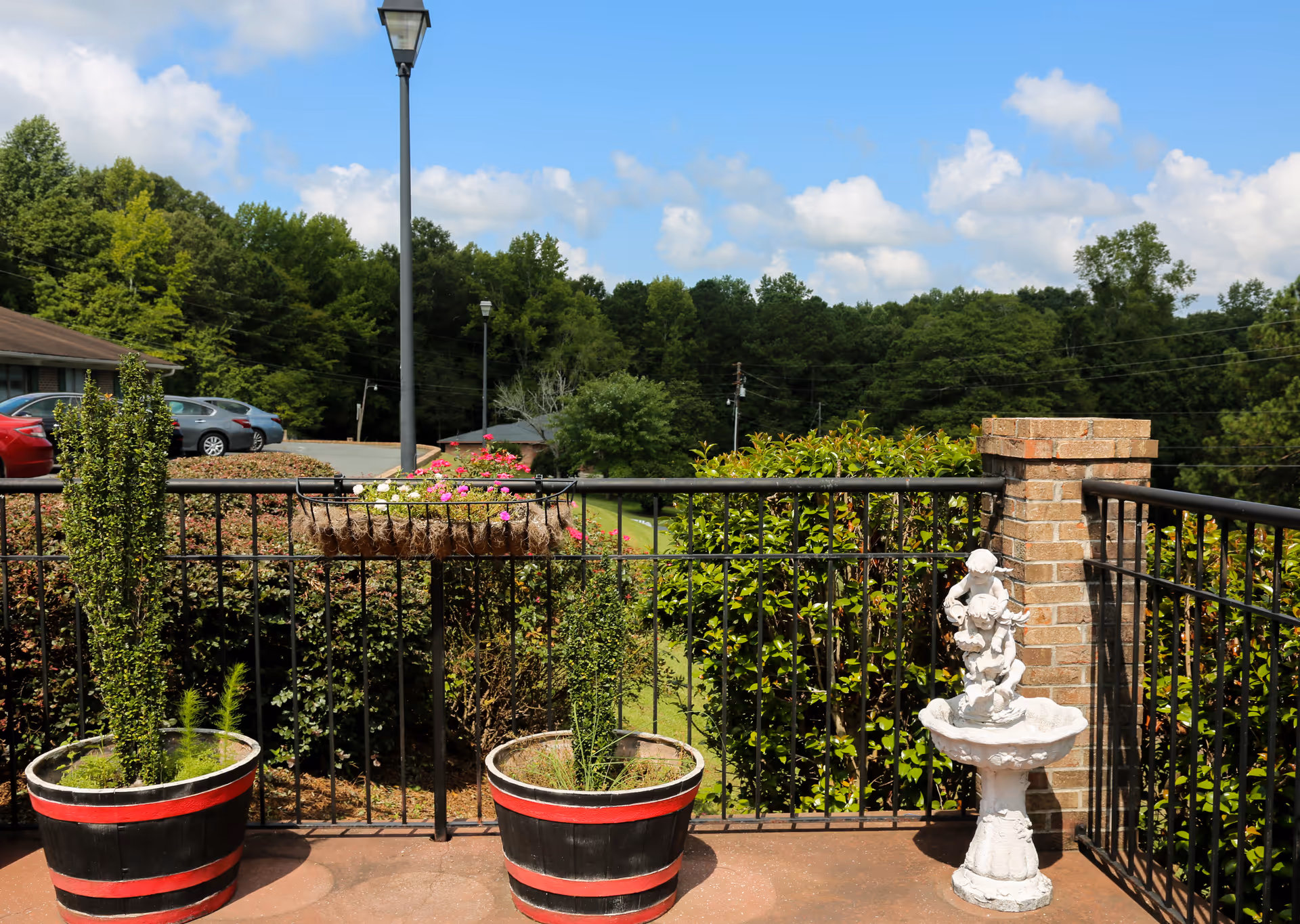 Outdoor patio area with black metal railing, two large black and red planters with greenery, a hanging basket with pink and white flowers, and a white decorative birdbath sculpture. In the background, there are green bushes, trees, a parking lot with cars, and a blue sky with scattered clouds.