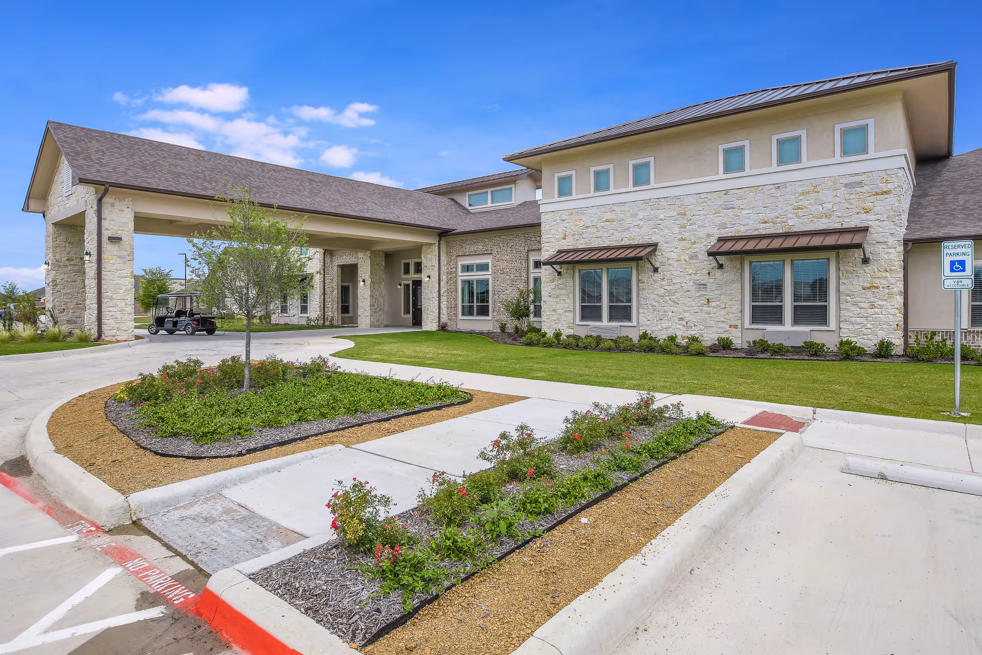 Exterior view of a senior living facility building with stone facade, a covered entrance driveway, landscaped flower beds, green lawn, and a reserved handicapped parking sign.