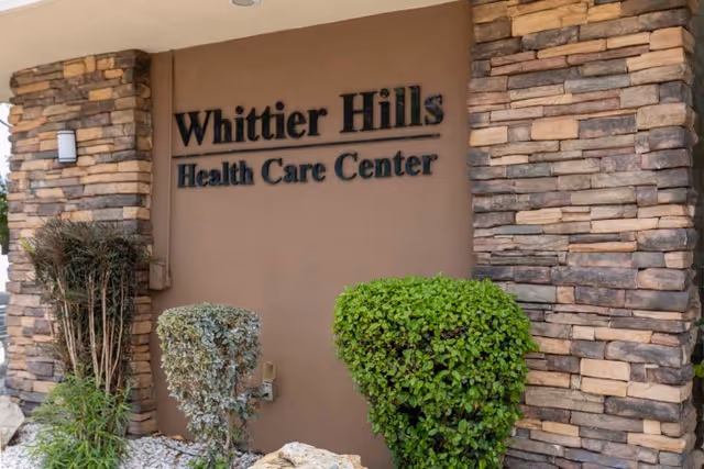 Exterior entrance wall with stone columns, landscaping, and a sign reading "Whittier Hills Health Care Center".