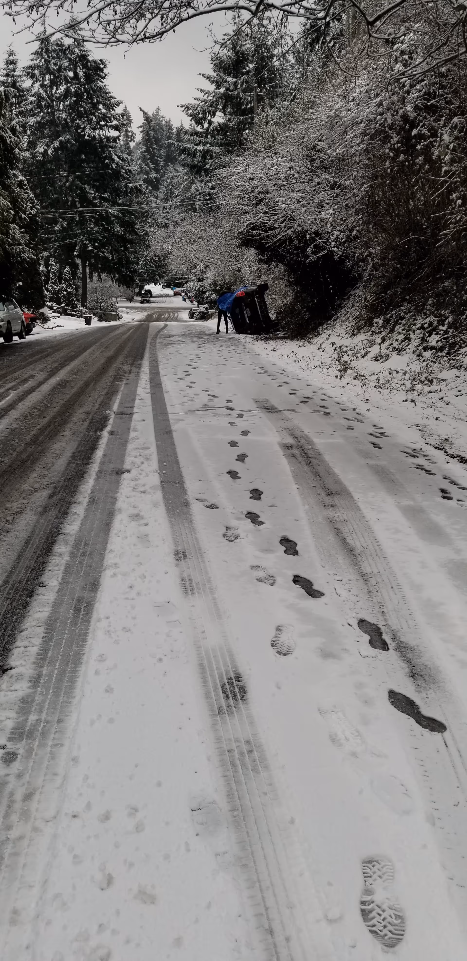 Snow-covered residential street with footprints, trees, and a person near an overturned vehicle.