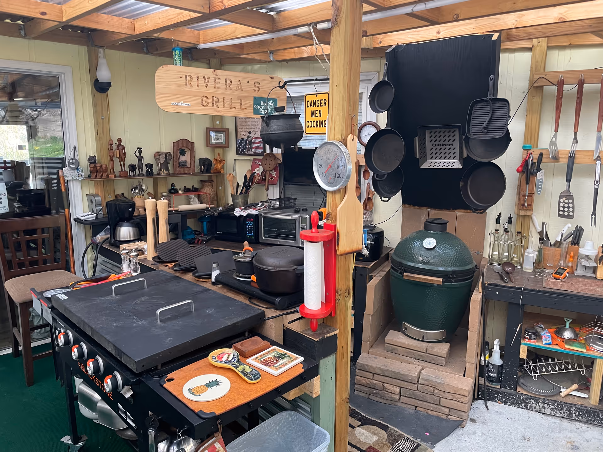 Outdoor kitchen area with grill, utensils, pans, and shelves beneath a wooden pergola.