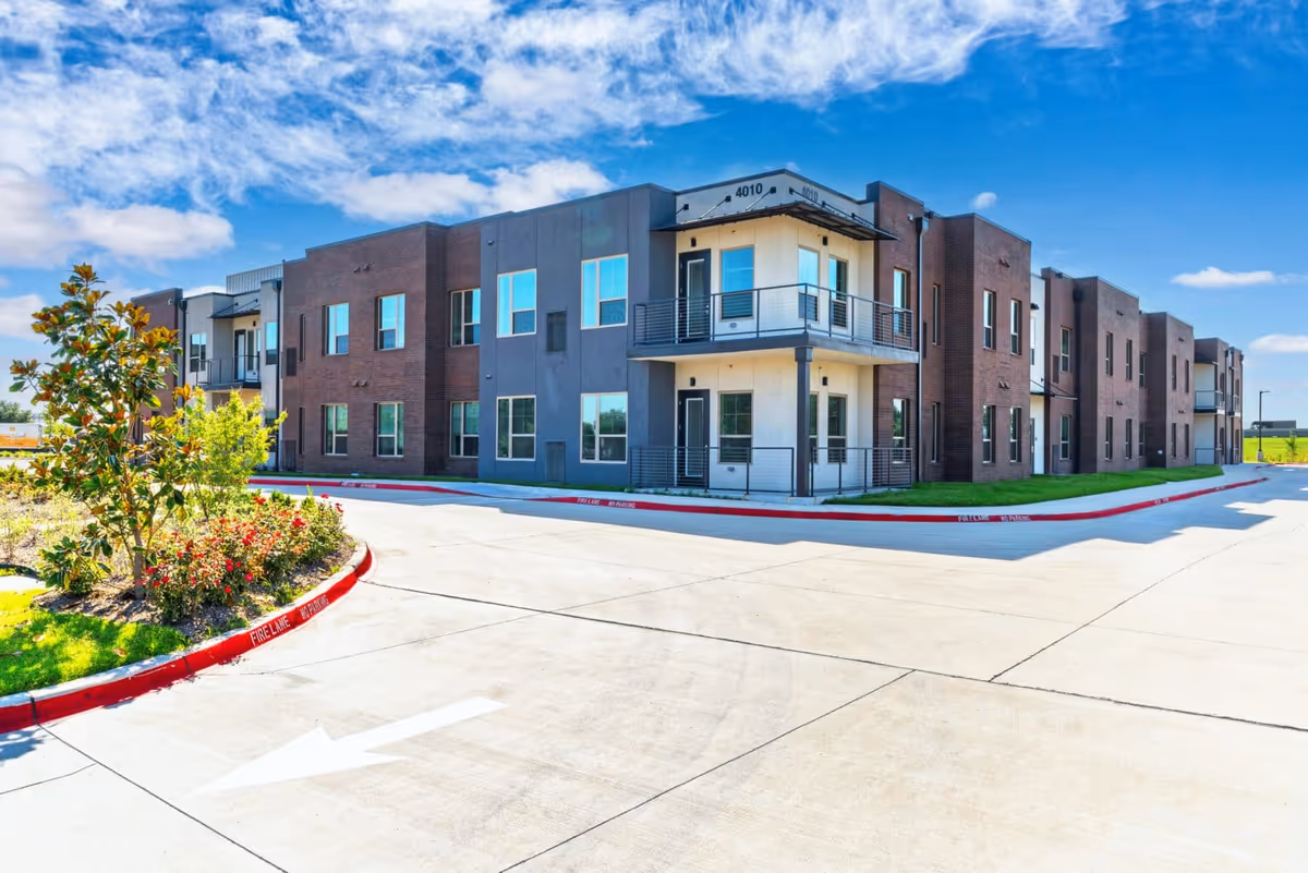 Exterior front view of a modern two-story brick and stucco senior living building with balconies, landscaping, and a driveway.