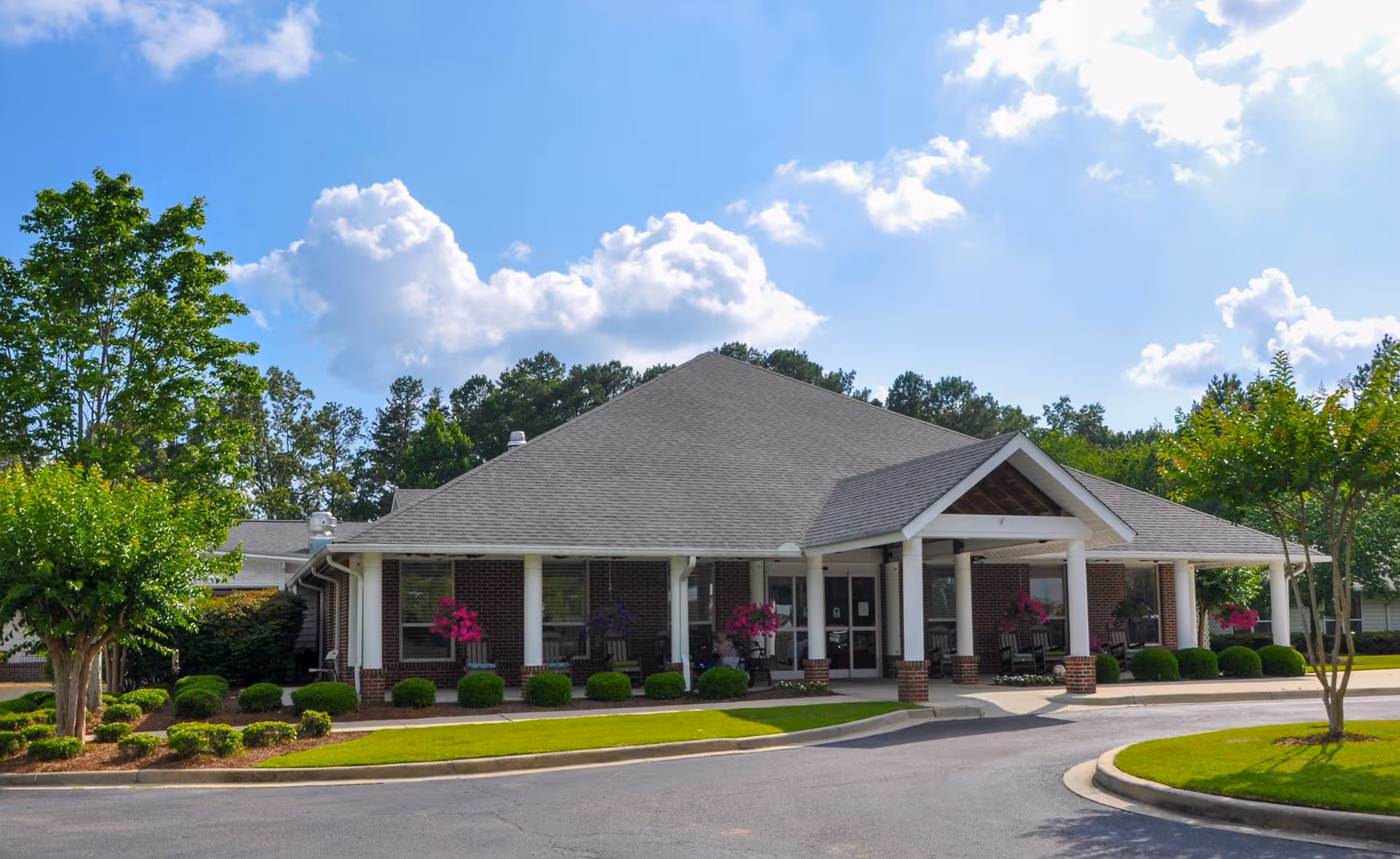 Front exterior view of a single-story brick building with a gray shingled roof, white columns, and a covered entrance. The building is surrounded by neatly trimmed bushes, small trees, and a well-maintained lawn under a partly cloudy blue sky.