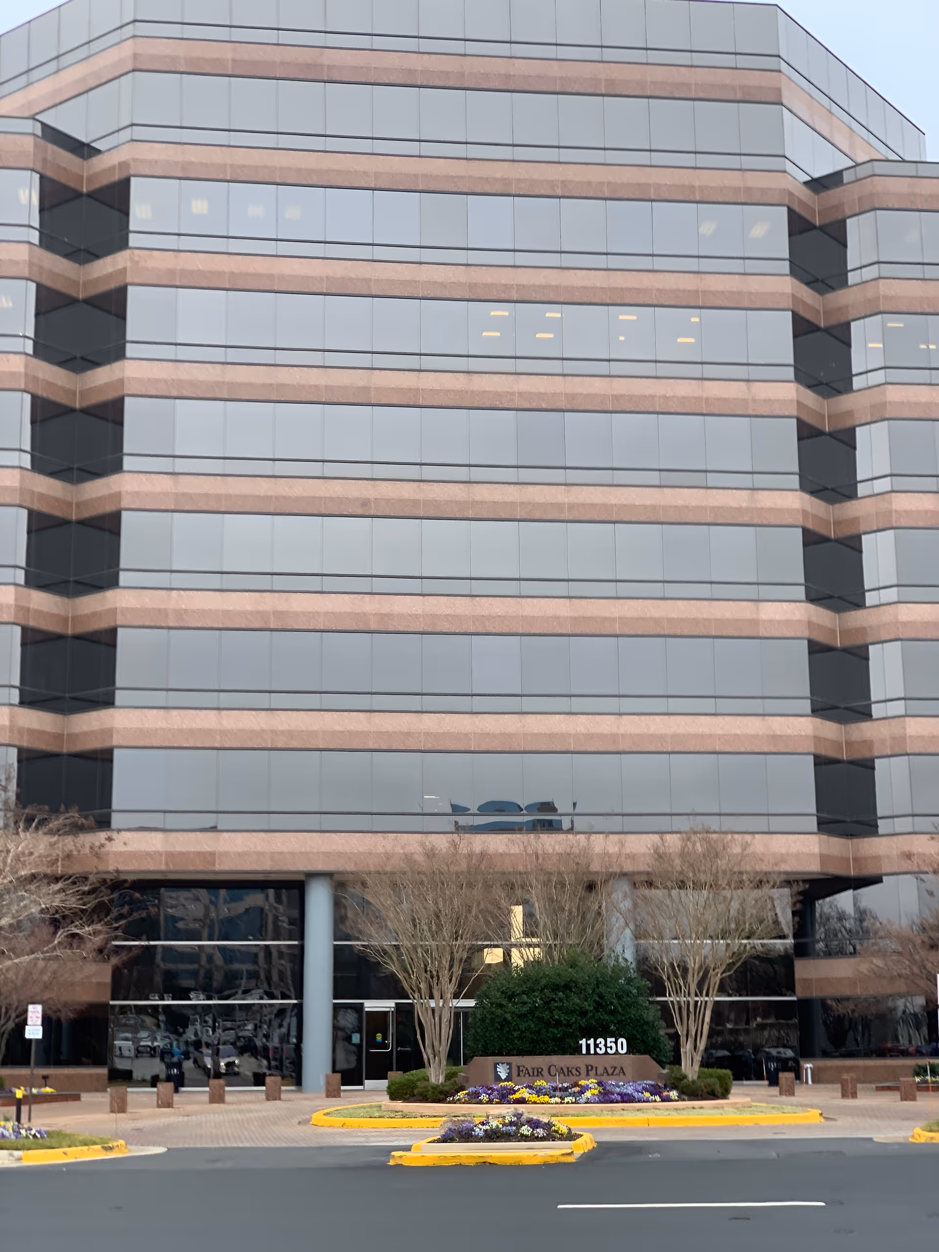Front exterior of a tall glass building with brick stripes and landscaping.