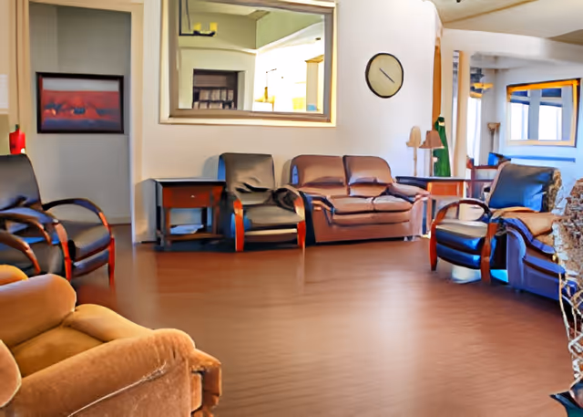 Spacious common living room with several armchairs and sofas arranged on a hardwood floor beneath a large mirror and wall clock.